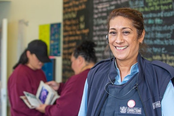 Corrections Officer stands smiling at camera with blackboard with te reo written on in the background